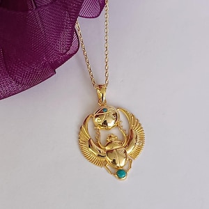 May include: Gold-colored pendant necklace featuring an Egyptian scarab beetle design with turquoise accents. The pendant has detailed wings and an eye symbol above the beetle. The necklace is on a delicate chain.