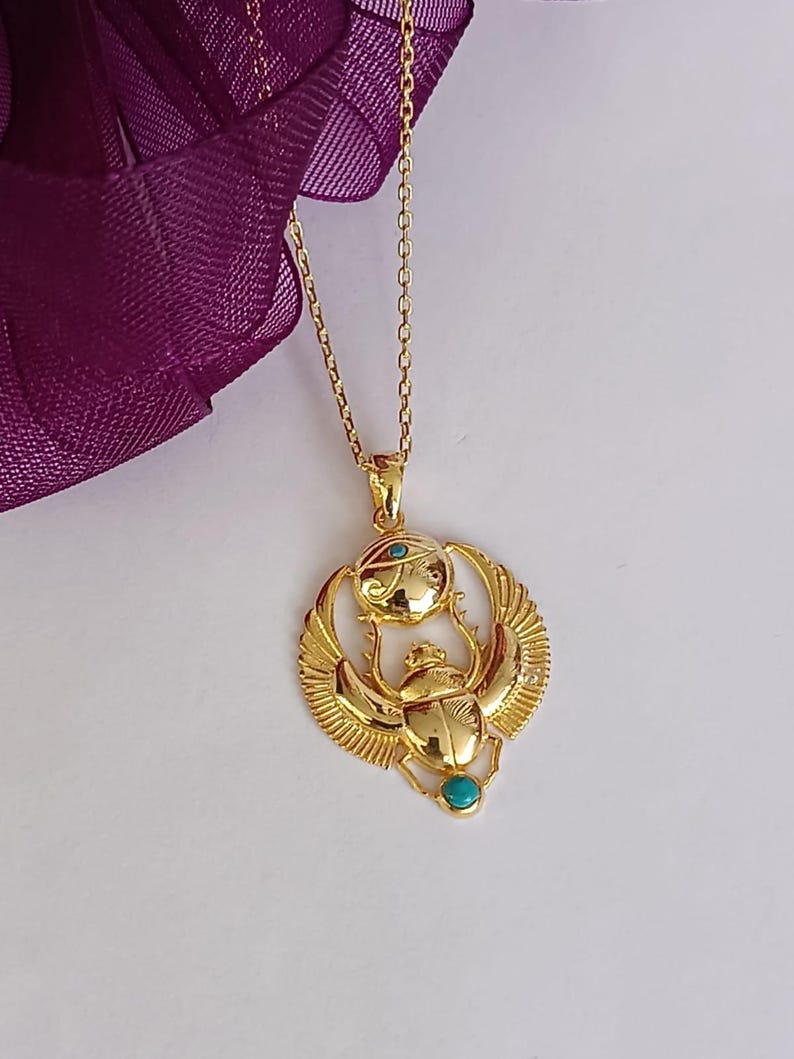 May include: Gold-colored pendant necklace featuring an Egyptian scarab beetle design with turquoise accents. The pendant has detailed wings and an eye symbol above the beetle. The necklace is on a delicate chain.