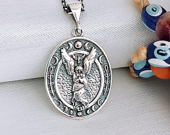 May include: Silver pendant necklace featuring an oval charm with an angel holding a cross. The charm is surrounded by a decorative border with crescent moons and leaves. The necklace is on a silver chain.