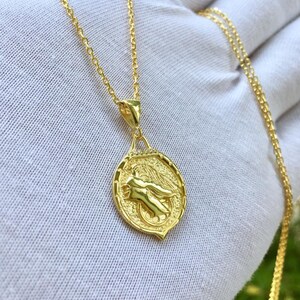 K&ouml;nnte beinhalten: Eine goldene Kettenkette mit einem Anh&auml;nger, der eine gefl&uuml;gelte Figur in einem kreisf&ouml;rmigen Design zeigt. Der Anh&auml;nger hat eine strukturierte Oberfl&auml;che und einen kleinen diamantartigen Stein oben.