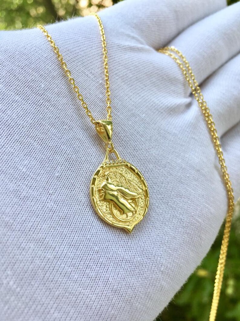 K&ouml;nnte beinhalten: Eine goldene Kettenkette mit einem Anh&auml;nger, der eine gefl&uuml;gelte Figur in einem kreisf&ouml;rmigen Design zeigt. Der Anh&auml;nger hat eine strukturierte Oberfl&auml;che und einen kleinen diamantartigen Stein oben.