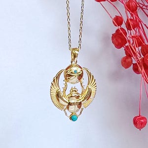 May include: Gold-colored pendant necklace featuring an Egyptian scarab beetle design. The pendant has a turquoise-colored stone and an eye symbol. The necklace is on a delicate gold chain, with red floral accents in the background.