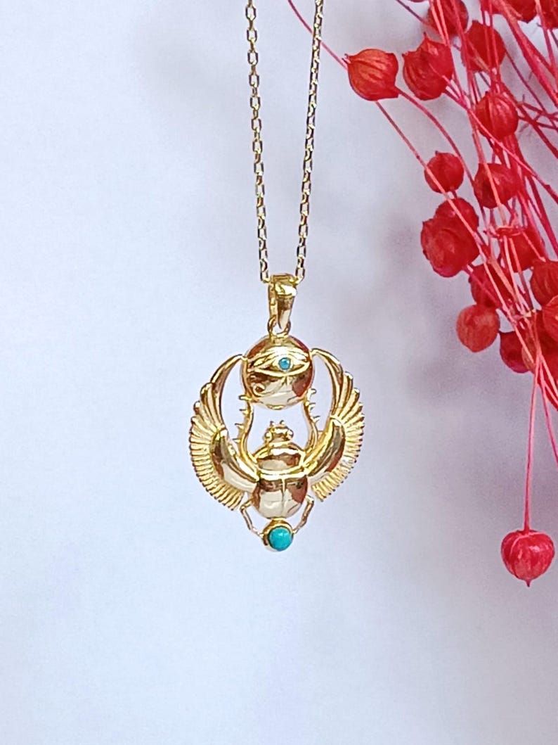 May include: Gold-colored pendant necklace featuring an Egyptian scarab beetle design. The pendant has a turquoise-colored stone and an eye symbol. The necklace is on a delicate gold chain, with red floral accents in the background.