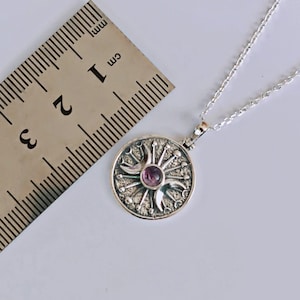 May include: A silver pendant necklace featuring a circular pendant with a sunburst design and a central purple gemstone. The pendant is approximately 1 cm in diameter and hangs on a delicate silver chain. The necklace is displayed on a white surface.