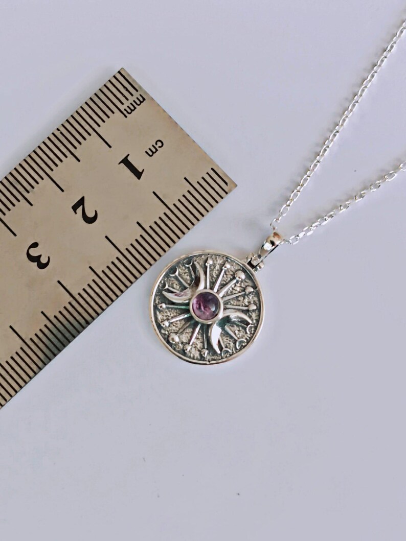 May include: A silver pendant necklace featuring a circular pendant with a sunburst design and a central purple gemstone. The pendant is approximately 1 cm in diameter and hangs on a delicate silver chain. The necklace is displayed on a white surface.