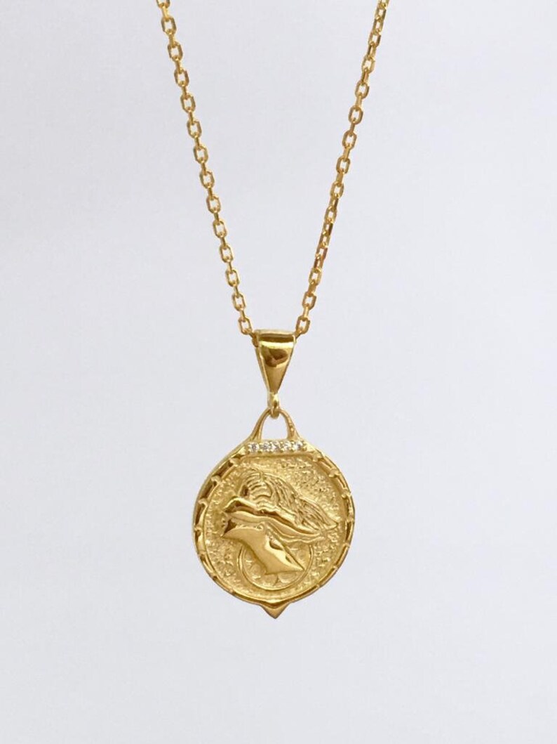 K&ouml;nnte beinhalten: Eine goldene Kettenkette mit einem runden Anh&auml;nger, der ein detailliertes Profil eines Frauenkopfes mit einer Krone zeigt. Der Anh&auml;nger ist mit kleinen, klaren Steinen verziert.