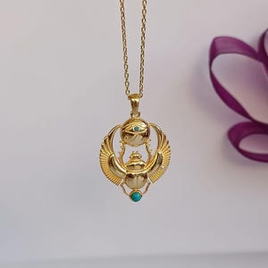 May include: A gold-colored pendant necklace featuring an Egyptian scarab beetle design. The pendant has turquoise accents and is suspended from a delicate chain. The wings of the beetle are detailed, and the overall design is ornate.