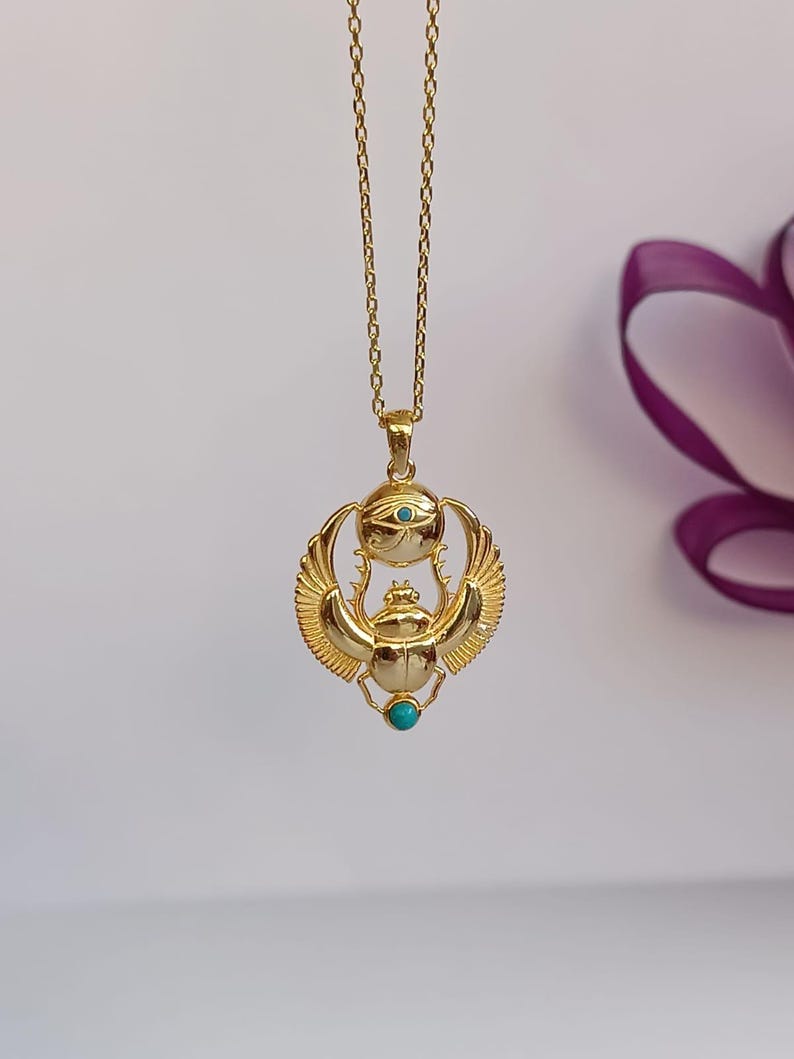 May include: A gold-colored pendant necklace featuring an Egyptian scarab beetle design. The pendant has turquoise accents and is suspended from a delicate chain. The wings of the beetle are detailed, and the overall design is ornate.