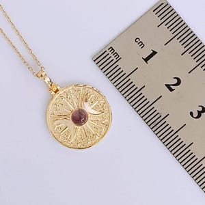 May include: A gold-colored pendant necklace with a circular pendant. The pendant features an intricate design with a small, round, pink gemstone in the center. The necklace is shown with a ruler, and the pendant is approximately 0.75 inches in diameter.