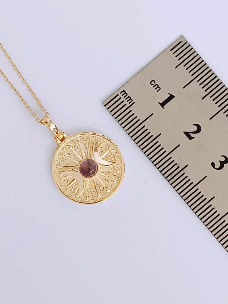 May include: A gold-colored pendant necklace with a circular pendant. The pendant features an intricate design with a small, round, pink gemstone in the center. The necklace is shown with a ruler, and the pendant is approximately 0.75 inches in diameter.