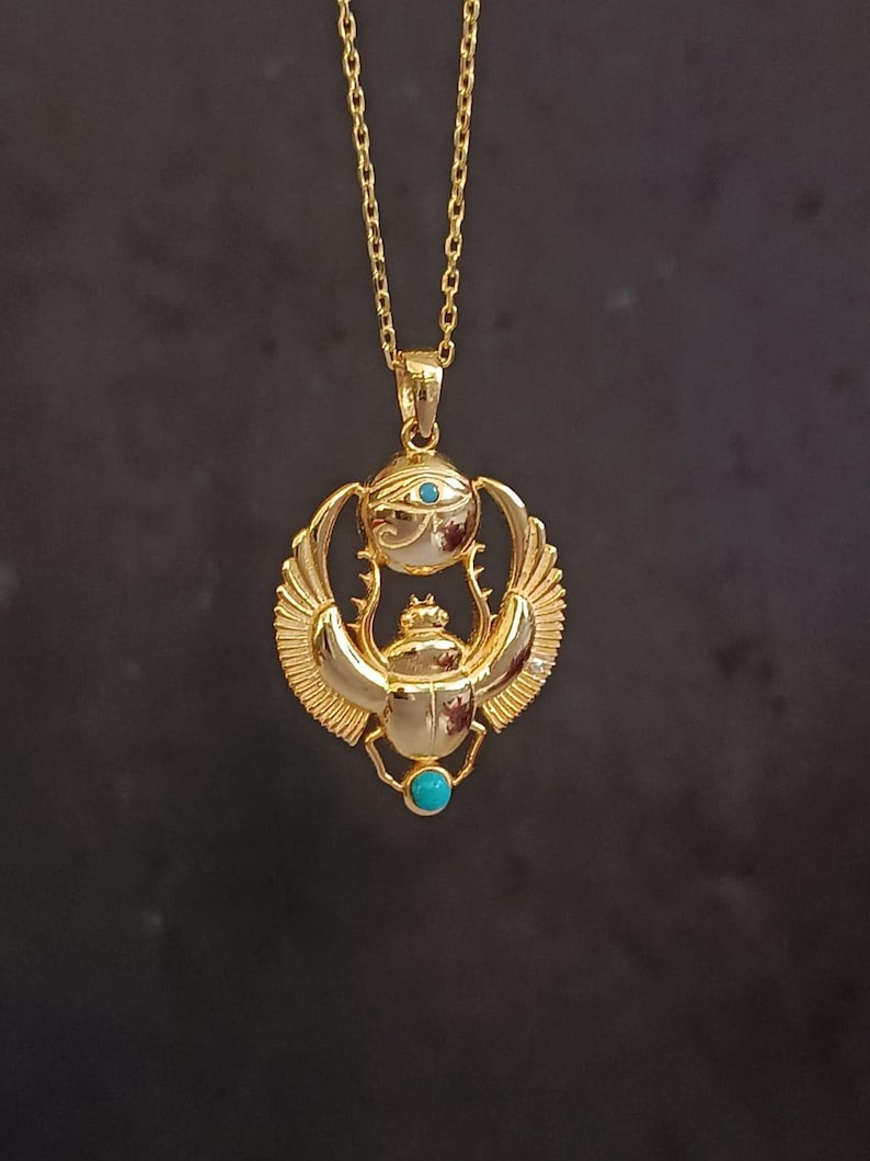 May include: Gold-colored pendant necklace featuring an Egyptian scarab beetle design. The pendant has a turquoise stone and an eye symbol. The necklace is on a gold chain.