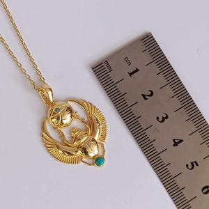 May include: A gold-colored pendant necklace featuring an Egyptian scarab beetle design with turquoise and clear stone accents. The pendant is approximately 1 inch in diameter and hangs from a delicate chain.