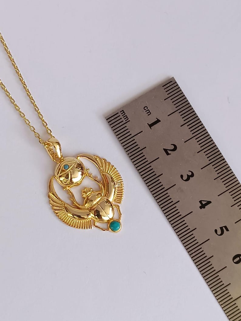 May include: A gold-colored pendant necklace featuring an Egyptian scarab beetle design with turquoise and clear stone accents. The pendant is approximately 1 inch in diameter and hangs from a delicate chain.