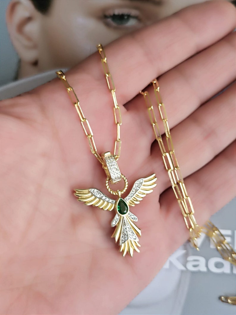 Pode incluir: Colar de corrente dourada com um pingente em forma de p&aacute;ssaro alado com uma gema verde e strass transparentes.