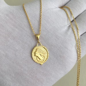 K&ouml;nnte beinhalten: Eine goldene Kettenkette mit einem Anh&auml;nger, der ein detailliertes Profil eines Frauenkopfes mit einer Krone zeigt. Der Anh&auml;nger ist von einem Kreis aus kleinen Diamanten umgeben.