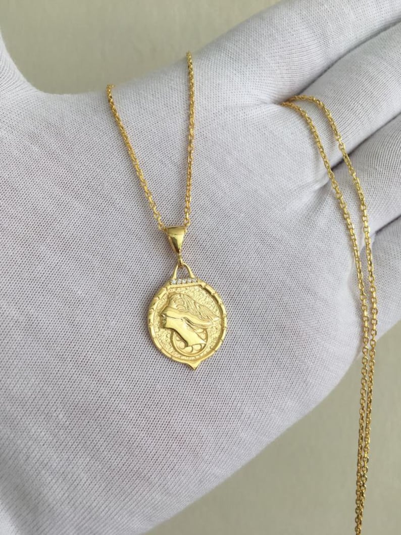 K&ouml;nnte beinhalten: Eine goldene Kettenkette mit einem Anh&auml;nger, der ein detailliertes Profil eines Frauenkopfes mit einer Krone zeigt. Der Anh&auml;nger ist von einem Kreis aus kleinen Diamanten umgeben.