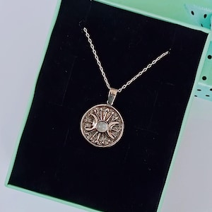 May include: A silver-toned necklace with a circular pendant featuring a sun and moon design. The pendant is set against a black background inside a light green gift box. The necklace chain is delicate and the pendant has intricate detailing.