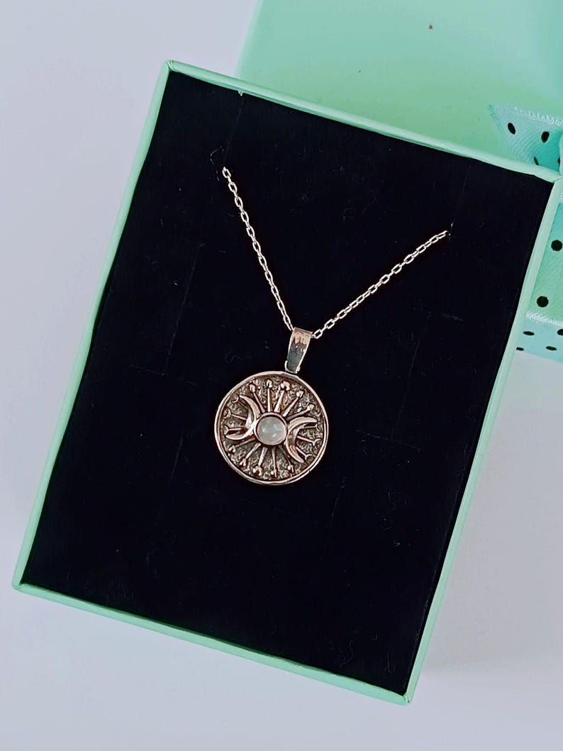 May include: A silver-toned necklace with a circular pendant featuring a sun and moon design. The pendant is set against a black background inside a light green gift box. The necklace chain is delicate and the pendant has intricate detailing.