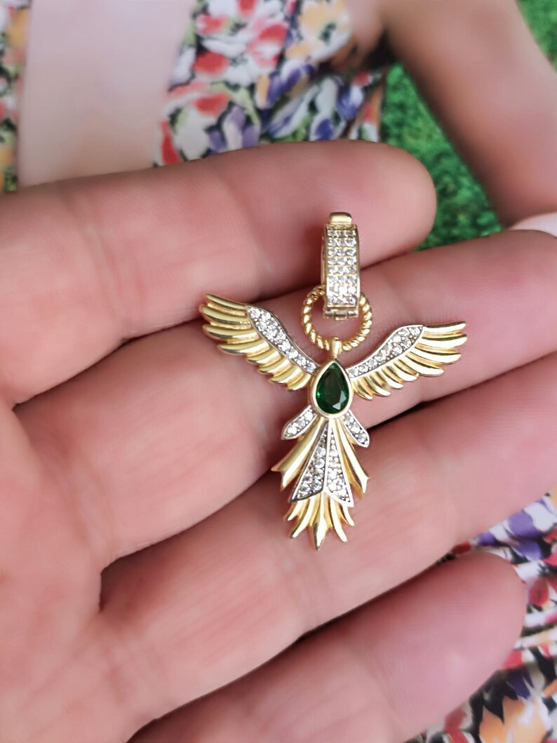 Pode incluir: Pingente de ouro com uma gema verde e strass transparentes em forma de um p&aacute;ssaro estilizado com as asas abertas.