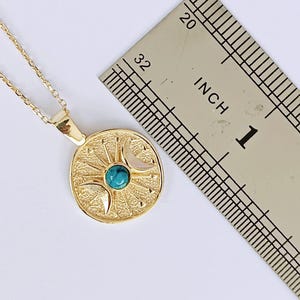May include: A gold-colored pendant necklace featuring a circular pendant with a sunburst design, a turquoise stone, and crescent moons. The pendant is approximately 1 inch in diameter. The necklace is on a white background.
