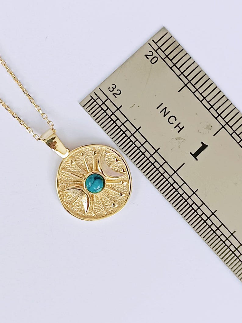 May include: A gold-colored pendant necklace featuring a circular pendant with a sunburst design, a turquoise stone, and crescent moons. The pendant is approximately 1 inch in diameter. The necklace is on a white background.