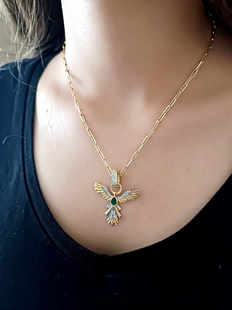 Pode incluir: Um colar de corrente de ouro com um pingente que apresenta um p&aacute;ssaro estilizado com as asas abertas. O p&aacute;ssaro tem uma gema verde no peito e &eacute; adornado com pequenas pedras transparentes.