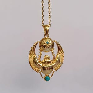 May include: A gold-colored pendant necklace featuring an Egyptian scarab beetle design. The pendant includes a turquoise-colored stone and an eye symbol. The necklace is suspended from a delicate gold chain.