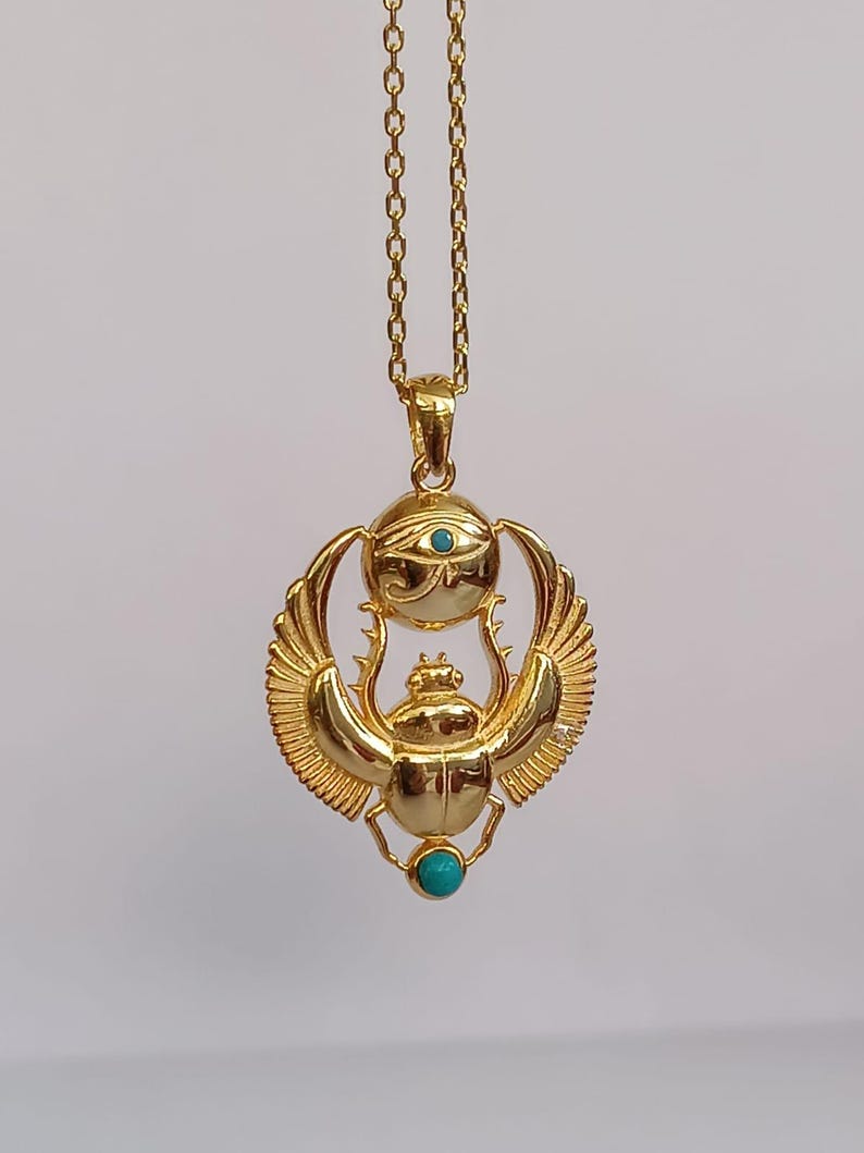 May include: A gold-colored pendant necklace featuring an Egyptian scarab beetle design. The pendant includes a turquoise-colored stone and an eye symbol. The necklace is suspended from a delicate gold chain.