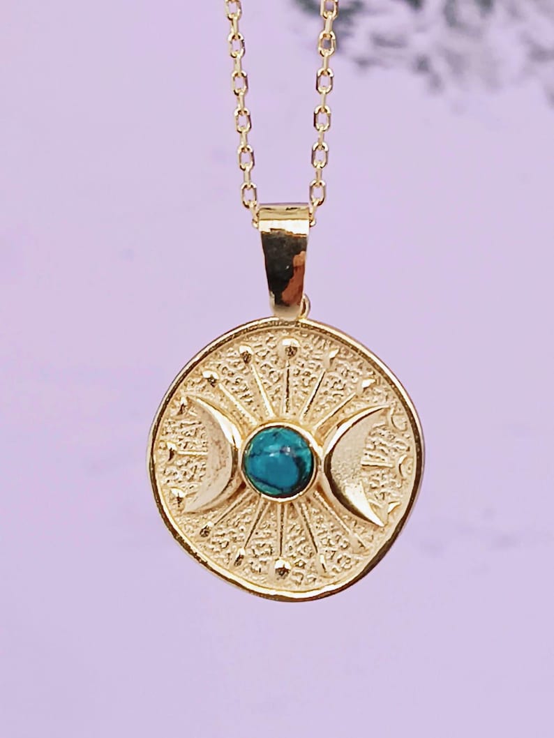 May include: Gold-colored pendant necklace featuring a circular pendant with a turquoise stone center and crescent moon designs. The pendant is suspended from a delicate chain. The necklace has a celestial or spiritual theme.