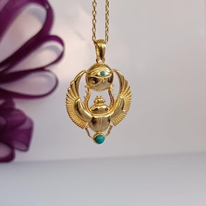 May include: Gold-colored pendant necklace featuring an Egyptian scarab beetle design with turquoise accents. The pendant includes the Eye of Horus and wings, suspended from a delicate chain. The necklace is displayed against a soft, neutral background.
