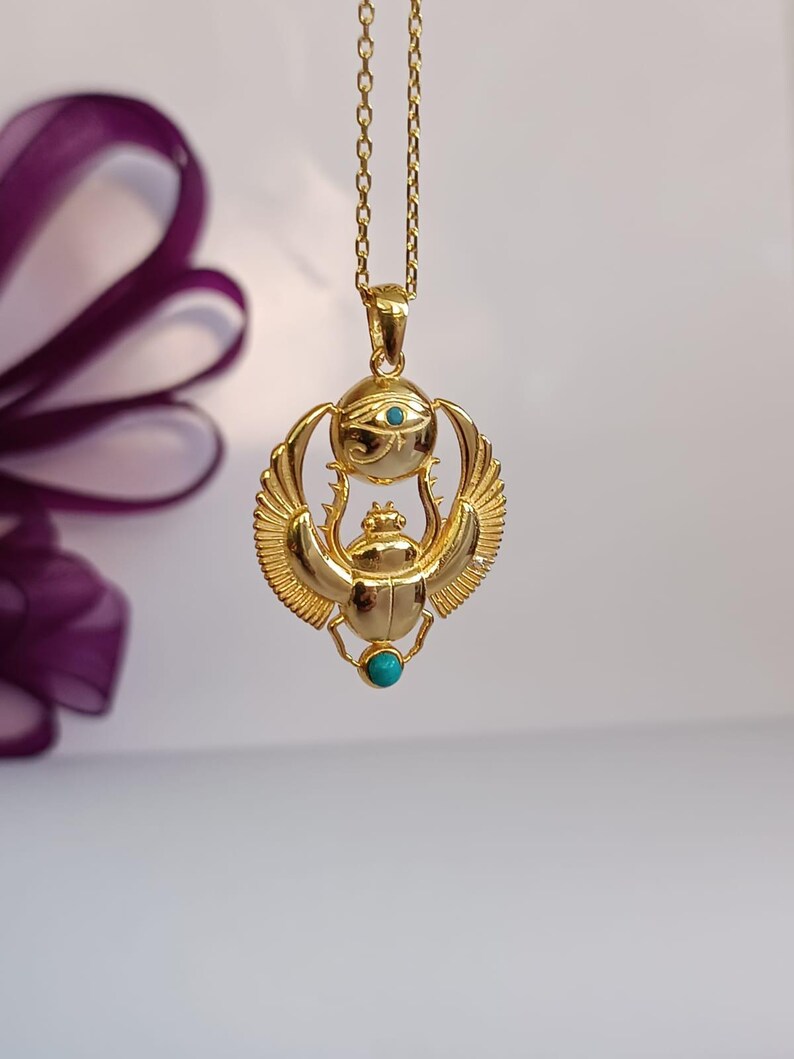 May include: Gold-colored pendant necklace featuring an Egyptian scarab beetle design with turquoise accents. The pendant includes the Eye of Horus and wings, suspended from a delicate chain. The necklace is displayed against a soft, neutral background.