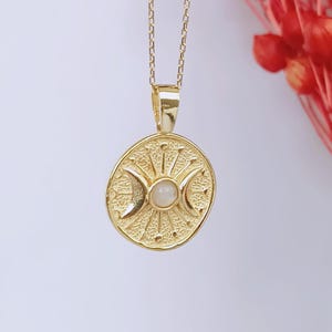 May include: Gold-colored pendant necklace featuring a circular pendant with a celestial design. The pendant has a central moonstone and crescent moon details. The necklace is suspended from a delicate chain, creating an elegant and mystical accessory.