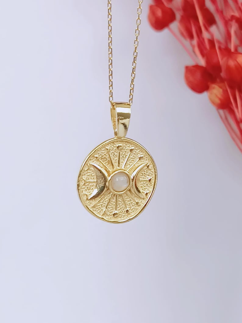 May include: Gold-colored pendant necklace featuring a circular pendant with a celestial design. The pendant has a central moonstone and crescent moon details. The necklace is suspended from a delicate chain, creating an elegant and mystical accessory.