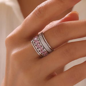 May include: A silver ring featuring a band of pink gemstones and a band of small, clear stones. The ring is on a finger, with a blurred background.