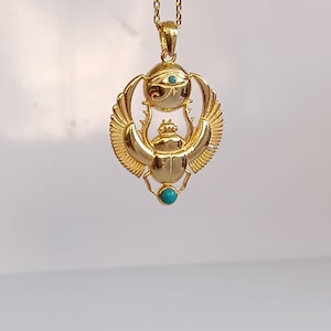 May include: A gold-colored pendant necklace featuring an Egyptian scarab beetle design. The pendant includes an eye of Horus and turquoise-colored accents. The necklace hangs from a delicate gold chain.