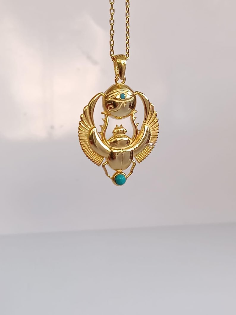 May include: A gold-colored pendant necklace featuring an Egyptian scarab beetle design. The pendant includes an eye of Horus and turquoise-colored accents. The necklace hangs from a delicate gold chain.