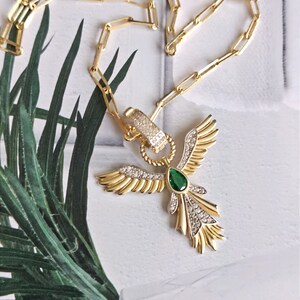 Pode incluir: Um colar de corrente de ouro com um pingente que apresenta um p&aacute;ssaro estilizado com as asas abertas. O p&aacute;ssaro tem uma gema verde no centro e &eacute; adornado com strass transparentes.