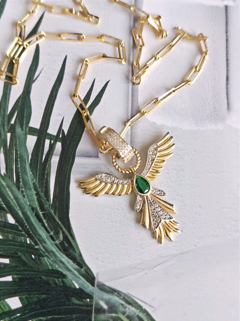 Pode incluir: Um colar de corrente de ouro com um pingente que apresenta um p&aacute;ssaro estilizado com as asas abertas. O p&aacute;ssaro tem uma gema verde no centro e &eacute; adornado com strass transparentes.