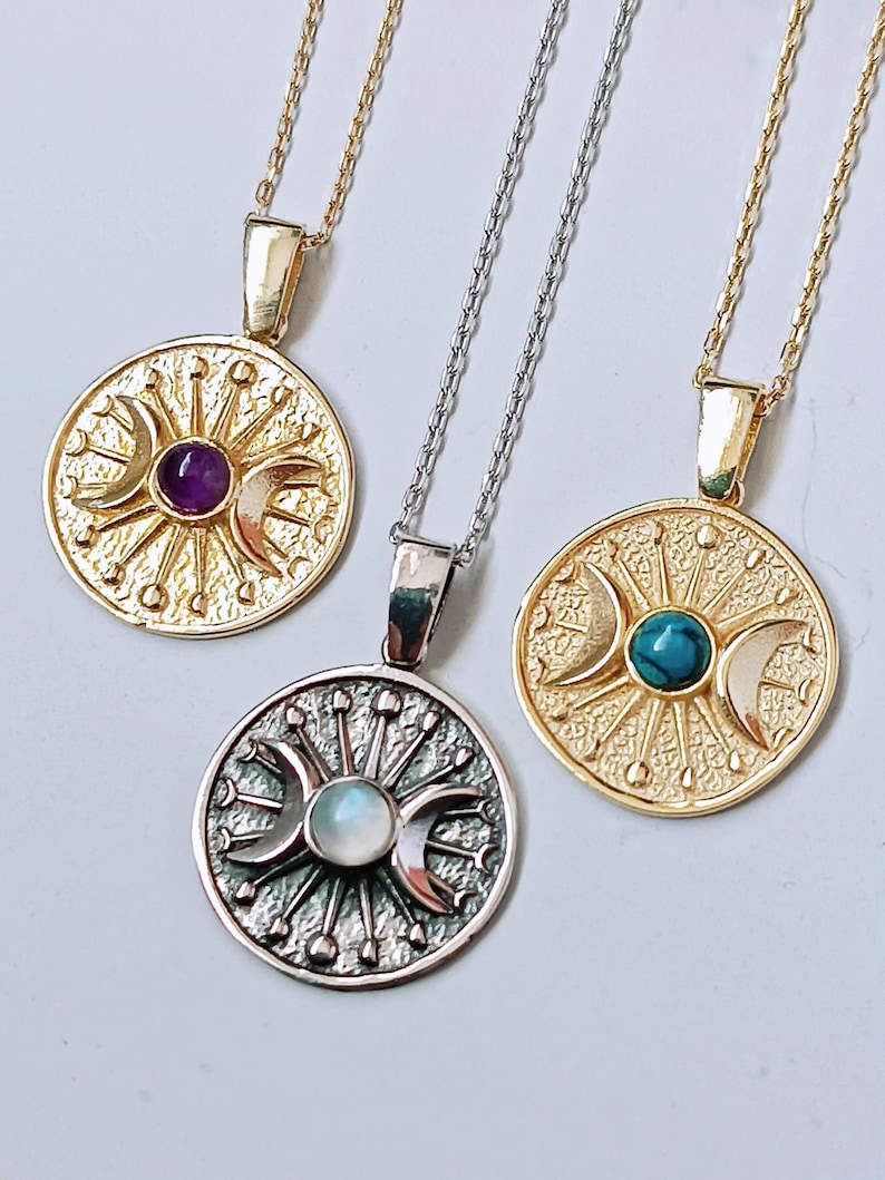 May include: Three necklaces with circular pendants. Two are gold-toned, one with an amethyst and the other with a turquoise stone. The third necklace is silver-toned with a moonstone. Each pendant features a sunburst design with crescent moons.
