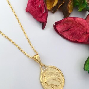 K&ouml;nnte beinhalten: Eine goldene Kettenkette mit einem Anh&auml;nger, der ein detailliertes Profil eines Frauenkopfes mit Fl&uuml;geln zeigt. Der Anh&auml;nger ist oval geformt und hat eine strukturierte Oberfl&auml;che. Die Kette ist eine zarte, kabelartige Kette.