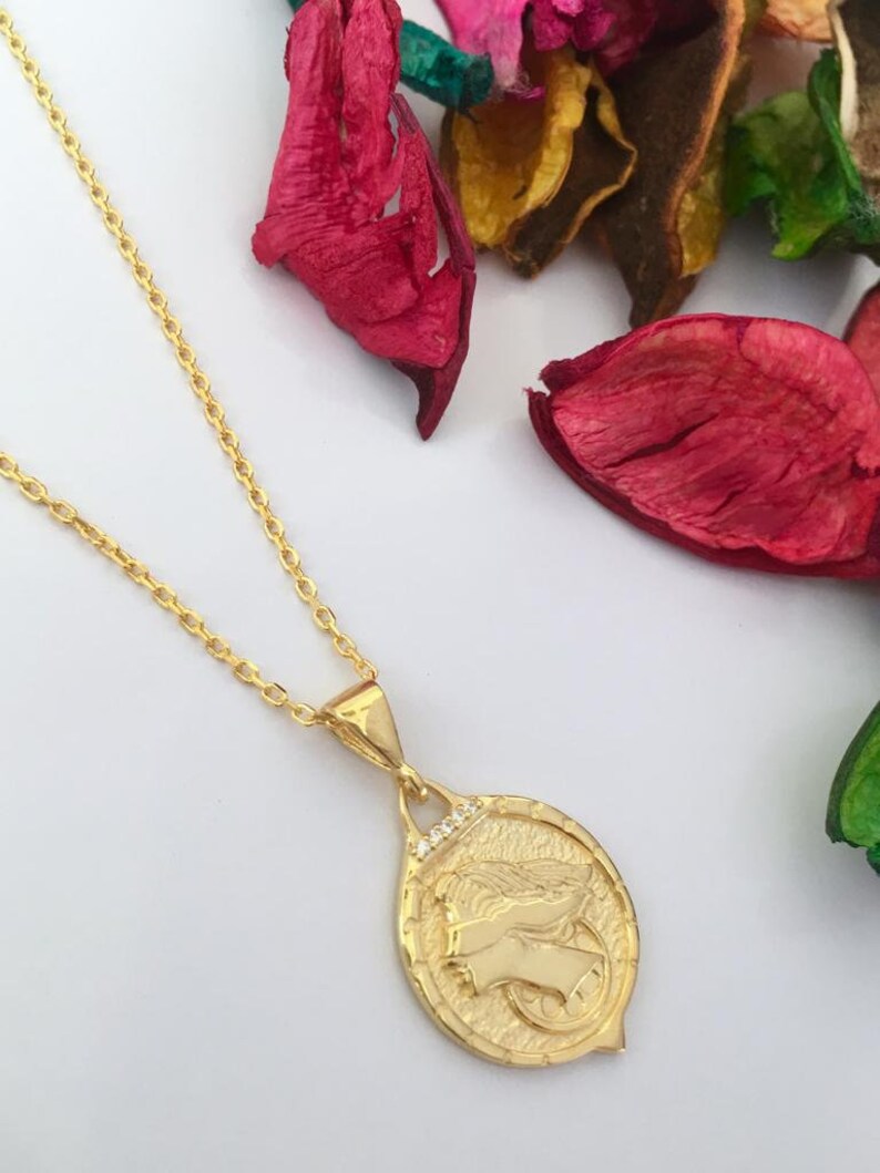 K&ouml;nnte beinhalten: Eine goldene Kettenkette mit einem Anh&auml;nger, der ein detailliertes Profil eines Frauenkopfes mit Fl&uuml;geln zeigt. Der Anh&auml;nger ist oval geformt und hat eine strukturierte Oberfl&auml;che. Die Kette ist eine zarte, kabelartige Kette.