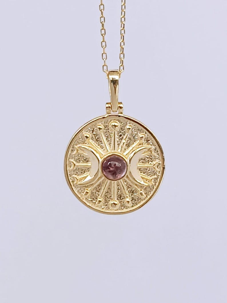 May include: A gold-colored pendant necklace featuring a circular pendant with a central pink gemstone. The pendant has a sunburst design with crescent moon details. The necklace is on a delicate chain.