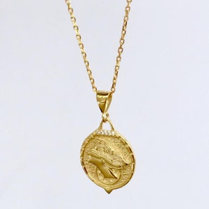 K&ouml;nnte beinhalten: Eine goldene Kettenkette mit einem Anh&auml;nger, der ein detailliertes Profil eines Frauenkopfes mit einer Krone zeigt. Der Anh&auml;nger ist von einem Kreis aus kleinen, klaren Steinen umgeben.