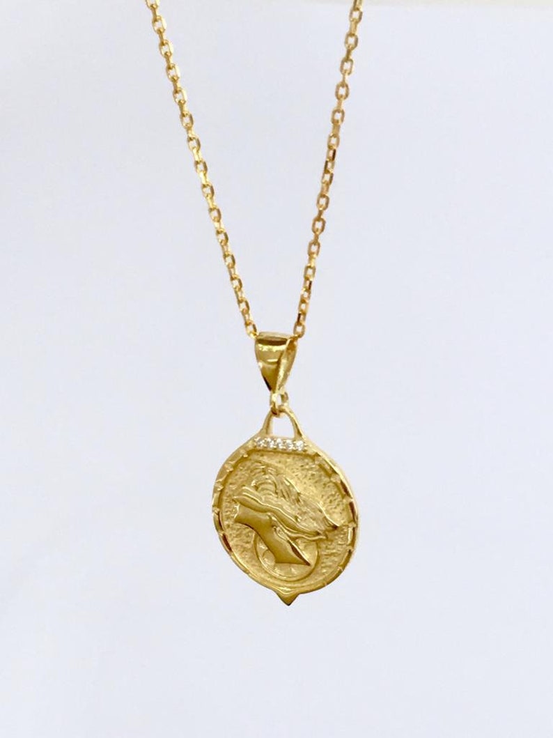 K&ouml;nnte beinhalten: Eine goldene Kettenkette mit einem Anh&auml;nger, der ein detailliertes Profil eines Frauenkopfes mit einer Krone zeigt. Der Anh&auml;nger ist von einem Kreis aus kleinen, klaren Steinen umgeben.