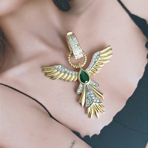 Pode incluir: Um colar dourado com um grande pingente que apresenta um p&aacute;ssaro estilizado com as asas abertas. O p&aacute;ssaro &eacute; adornado com strass transparentes e verdes.