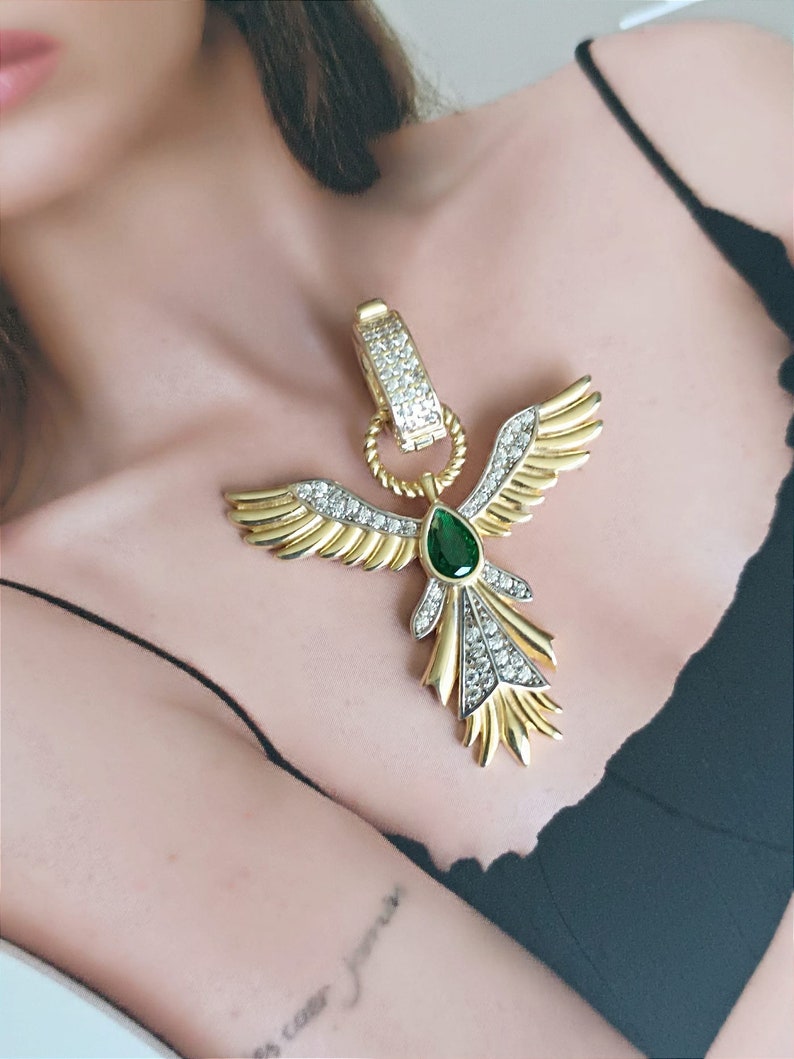 Pode incluir: Um colar dourado com um grande pingente que apresenta um p&aacute;ssaro estilizado com as asas abertas. O p&aacute;ssaro &eacute; adornado com strass transparentes e verdes.