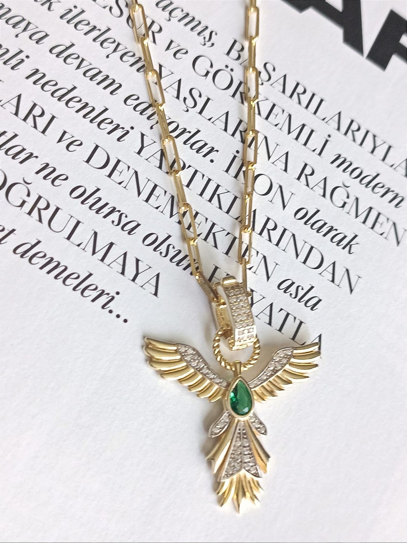 Pode incluir: Um colar de corrente de ouro com um pingente que apresenta um anjo alado com uma gema verde e detalhes de diamantes.