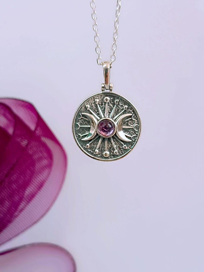 May include: A silver pendant necklace featuring a circular pendant with a central purple gemstone. The pendant has a sunburst design with crescent moons and star details. The necklace chain is also silver.