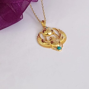May include: A gold-colored pendant necklace featuring an Egyptian scarab beetle design. The pendant has intricate details and small turquoise-colored accents. The necklace is on a delicate gold chain, with a purple fabric in the background.