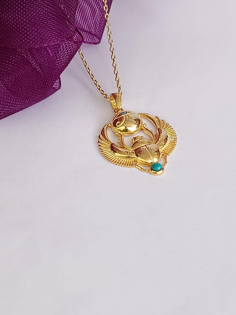 May include: A gold-colored pendant necklace featuring an Egyptian scarab beetle design. The pendant has intricate details and small turquoise-colored accents. The necklace is on a delicate gold chain, with a purple fabric in the background.